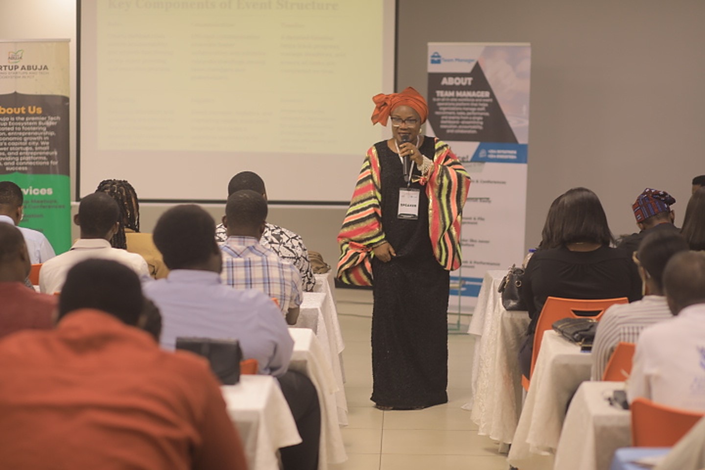 A speaker addresses a full room of founders seated at tables during a Startup Abuja session, with Startup Abuja and Team Manager banners behind her.