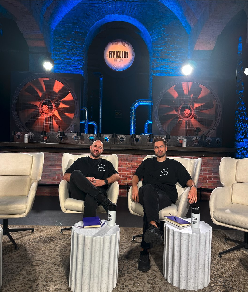 The two Callsy co-founders, in black Callsy T-shirts, seated in white armchairs on the set of Shark Tank Lithuania, with big orange-lit fans behind them.
