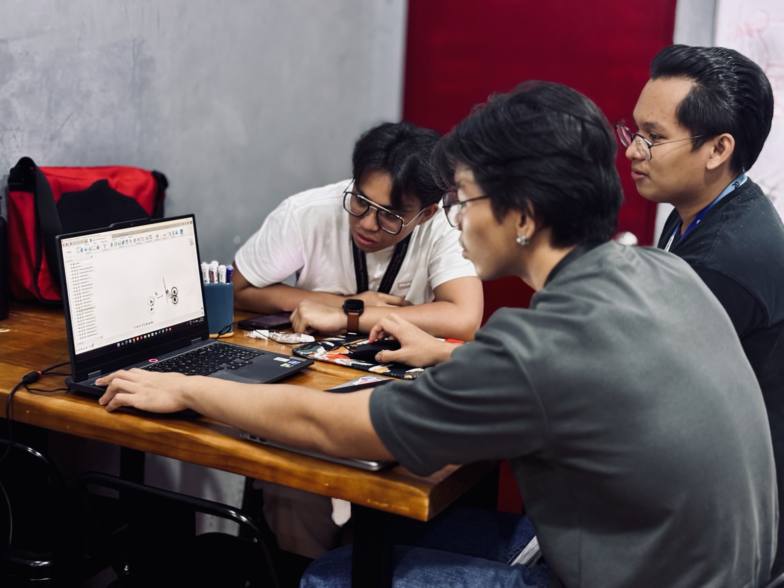 Three young engineers at a workshop table, one working on a laptop showing a CAD drawing while the others watch.